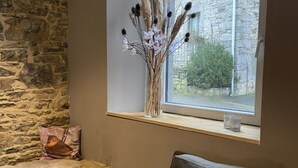 Et hyggeligt rum med en stenvæg, en polstret bænk, et vindue med udsigt over grønne omgivelser, og en vase med tørrede planter.