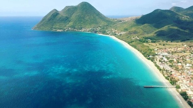An aerial view of a coastal area with a clear blue sea, a sandy beach, and a town with buildings. - Kay Agapi - on the beach - Diamant (Le Diamant)