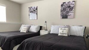 A bedroom with two beds, each with a black bedspread and checkered pillows, a bedside table with a lamp, and two wall-mounted artworks. - Ray's Place (Roy)