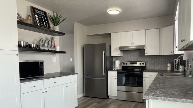 A modern kitchen with white cabinets, stainless steel appliances, and a wooden floor. - Ray's Place (Roy)