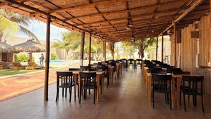 A covered outdoor dining area with wooden tables and chairs, a pool in the background, and a thatched roof structure.
