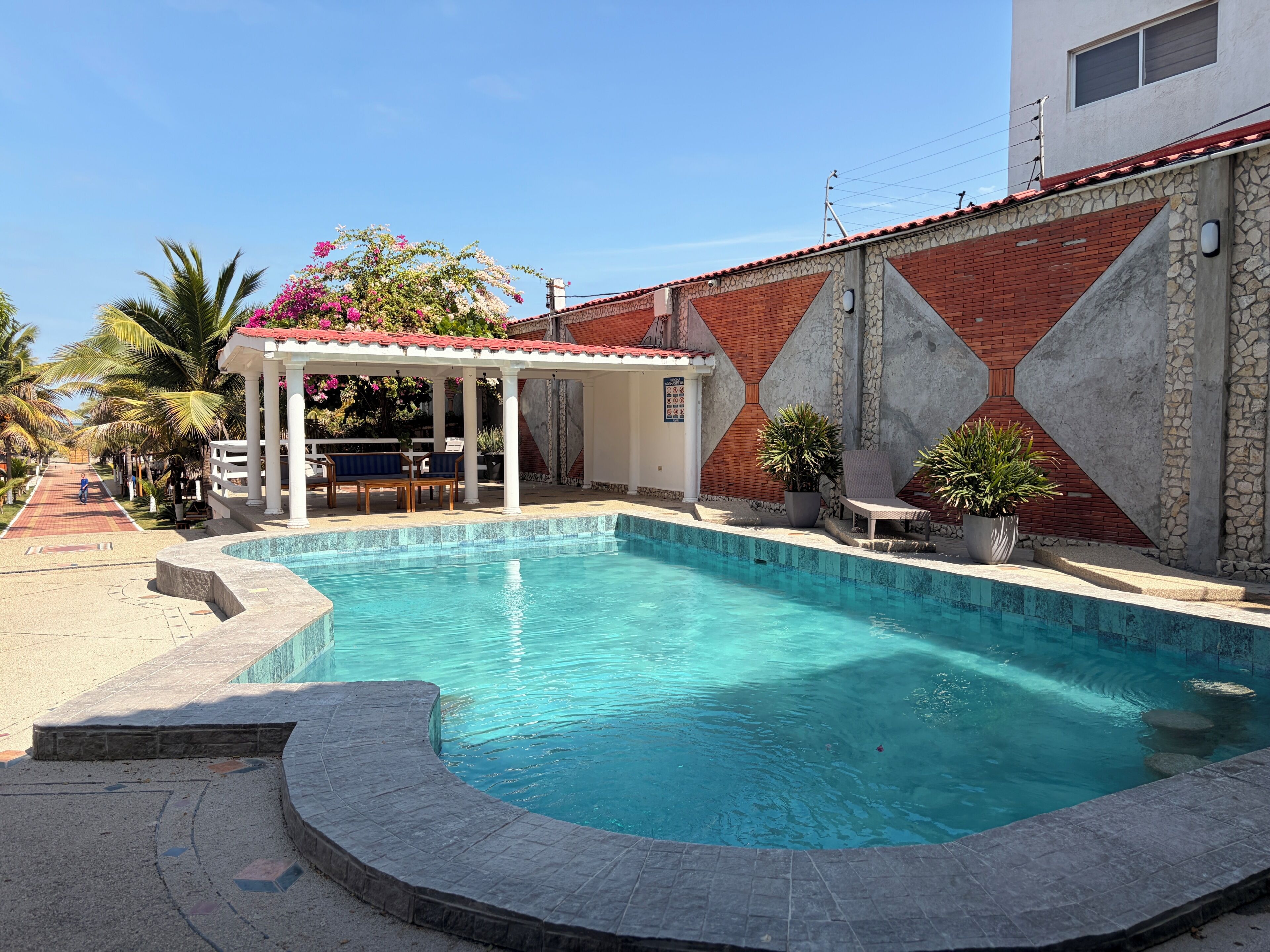 Una piscina con un pérgola, un patio con sillas y un muro de ladrillo con diseños geométricos.