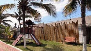 A playground with a slide and climbing structure, surrounded by palm trees and a bamboo fence.