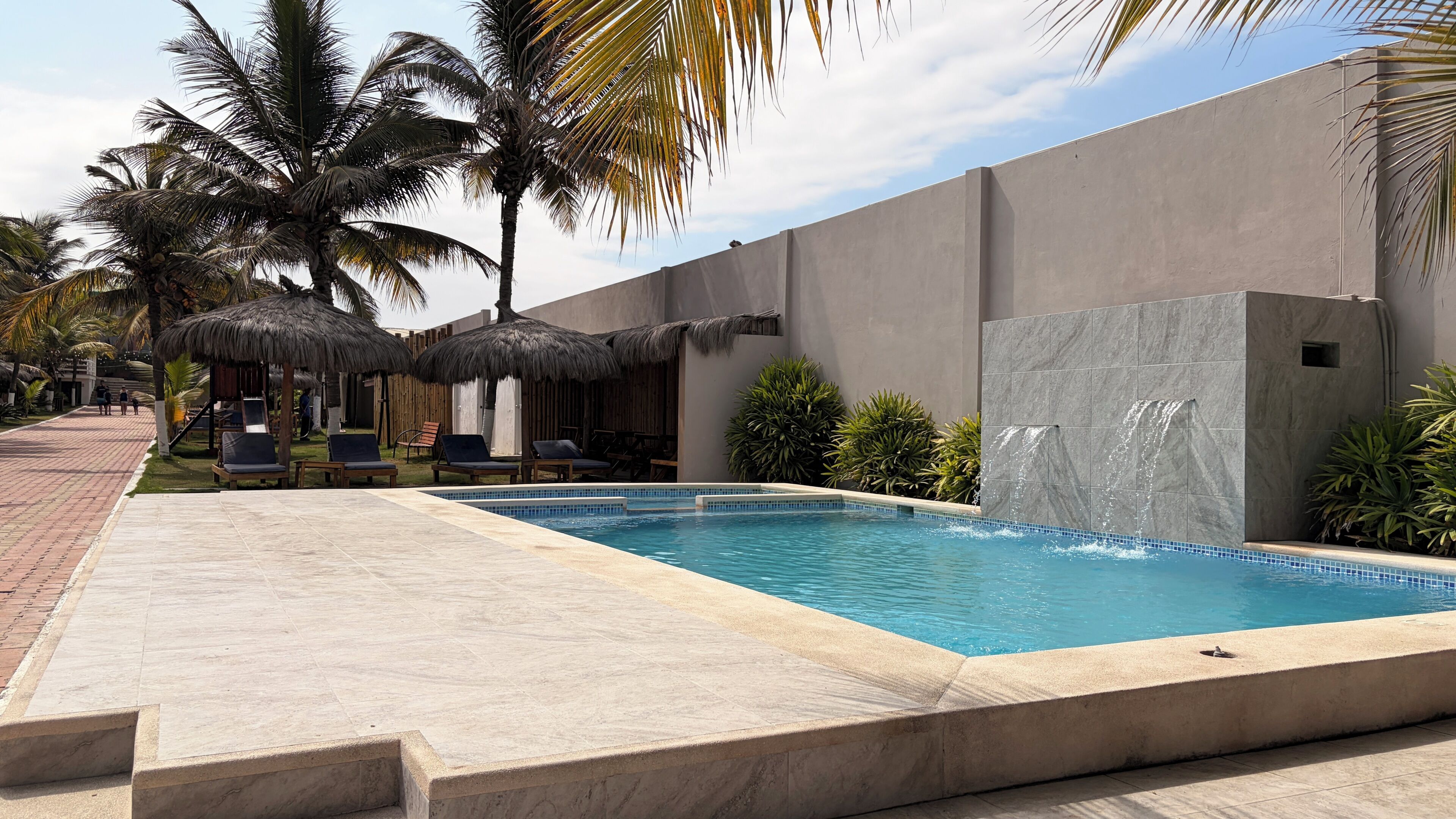 A swimming pool with a stone wall, palm trees, and lounge chairs.