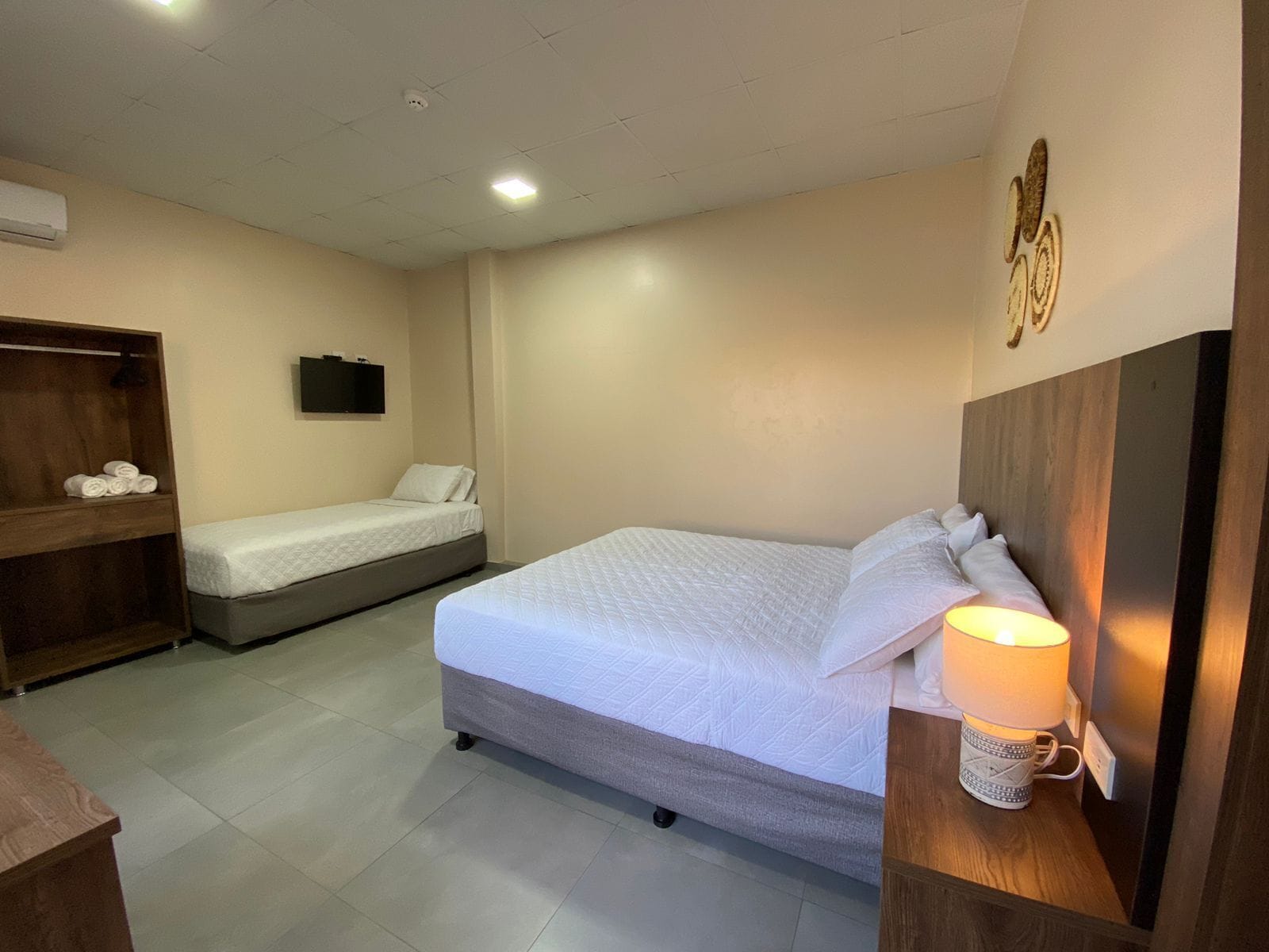 A hotel room with a large bed, a smaller bed, a wooden wardrobe, a wall-mounted TV, and a bedside table with a lamp.