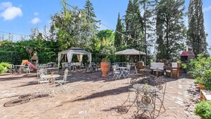 Un patio al aire libre con suelo de ladrillo, mobiliario de metal y madera, un pabellón y luces colgantes.