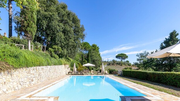 Una piscina rectangular con borde de piedra, rodeada por un patio de piedra y protegida por sombrillas.