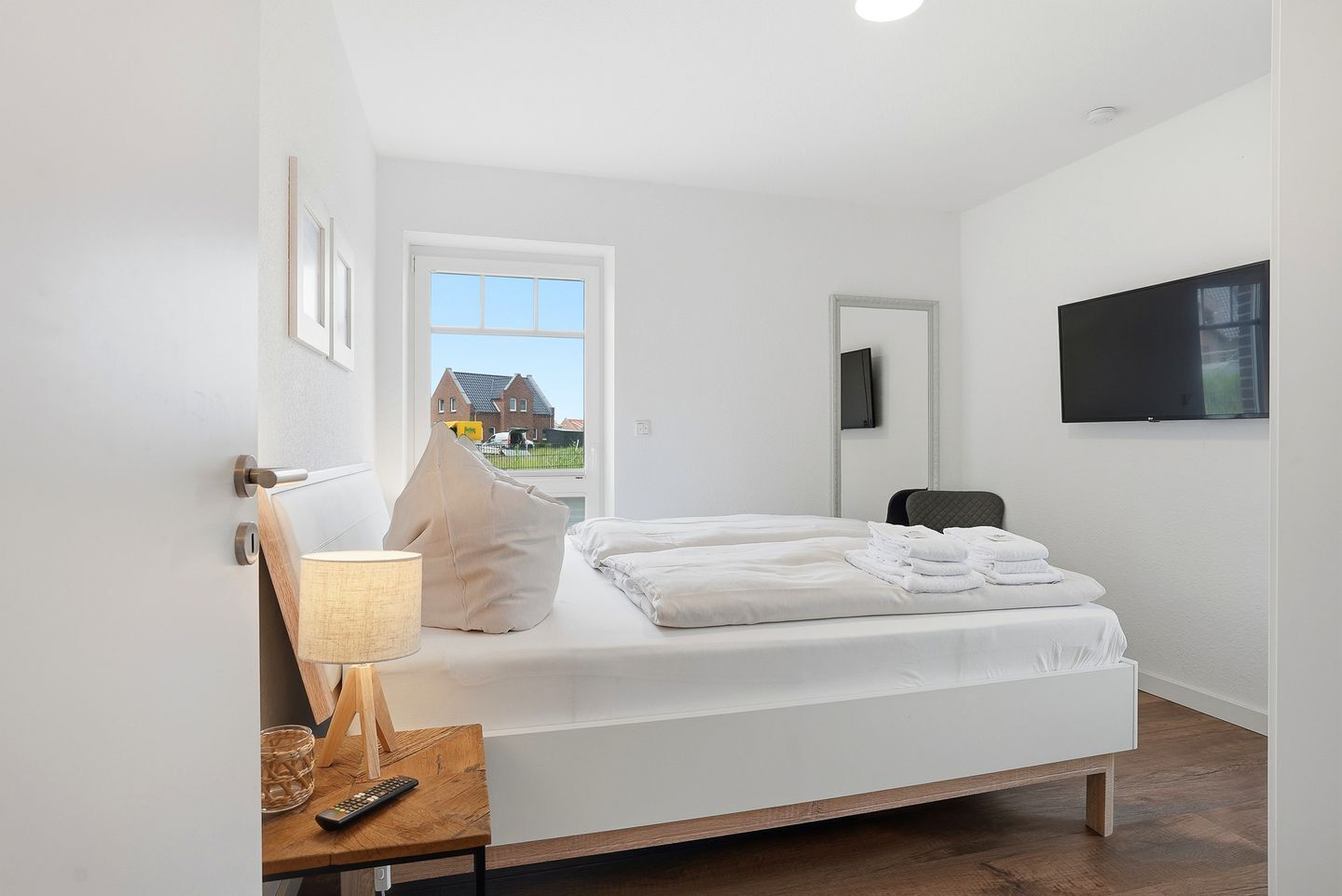 Un dormitorio con cama, una mesita de noche con lámpara, una silla, televisor montado en la pared y ventana con vistas a casas.