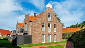 A modern brick building with a unique triangular roof design, multiple windows, and a paved pathway leading to the entrance. - Lieblingsort - Neubauwohnung mit Hund für bis zu 4 Personen im Greetsieler Grachtenpark (Krummhörn)