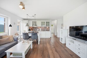 A modern kitchen with white cabinets, a dining area with grey chairs, and a TV on a white cabinet.