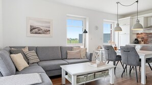 A modern living room with a grey sectional sofa, a white coffee table, and a dining area with grey chairs and a white table. - Lieblingsort - Neubauwohnung mit Hund für bis zu 4 Personen im Greetsieler Grachtenpark (Krummhörn)
