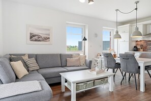A modern living room with a grey sectional sofa, a white coffee table, and a dining area with grey chairs and a white table. - Lieblingsort - Neubauwohnung mit Hund für bis zu 4 Personen im Greetsieler Grachtenpark (Krummhörn)