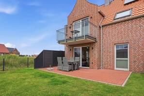 A brick house with a balcony, a fenced yard, and a patio with outdoor furniture. - Lieblingsort - Neubauwohnung mit Hund für bis zu 4 Personen im Greetsieler Grachtenpark (Krummhörn)