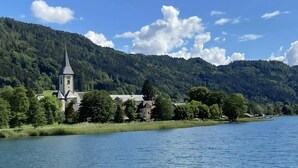 A church with a tall steeple by a lake, surrounded by lush greenery and mountains. - Holiday Home Bodensdorf Near Gerlitzen Ski Area (Steindorf am Ossiacher See)