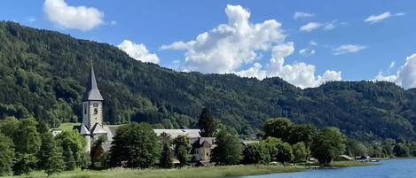 A church with a tall steeple by a lake, surrounded by lush greenery and mountains.