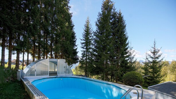 Ein Schwimmbad mit Glasüberdachung, umgeben von hohen Bäumen und blauem Himmel.