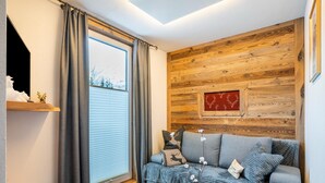 A cozy living room with a wooden wall, a gray sofa, a coffee table with a colorful abstract painting, and a window with curtains.