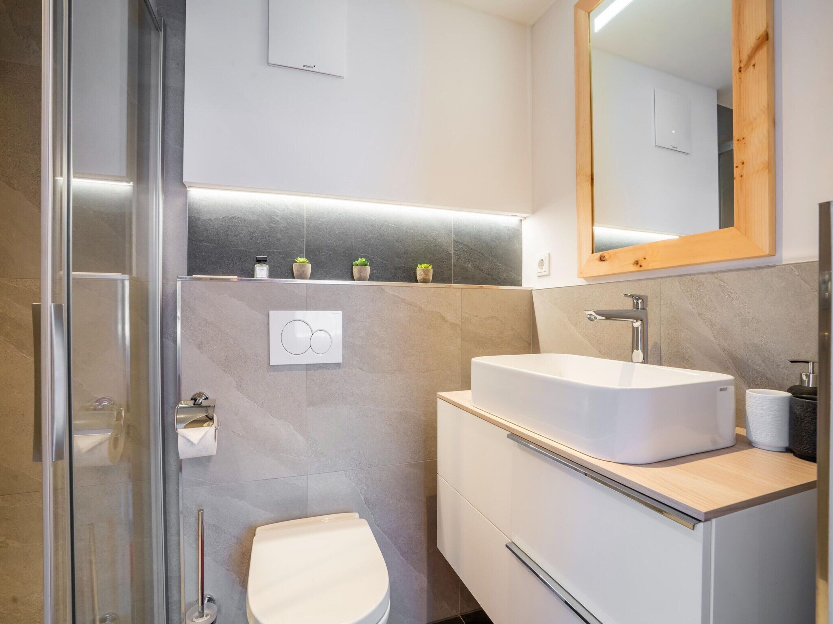 A modern bathroom with a glass shower enclosure, a white toilet, a large sink with a wooden countertop, and a mirror above the sink.