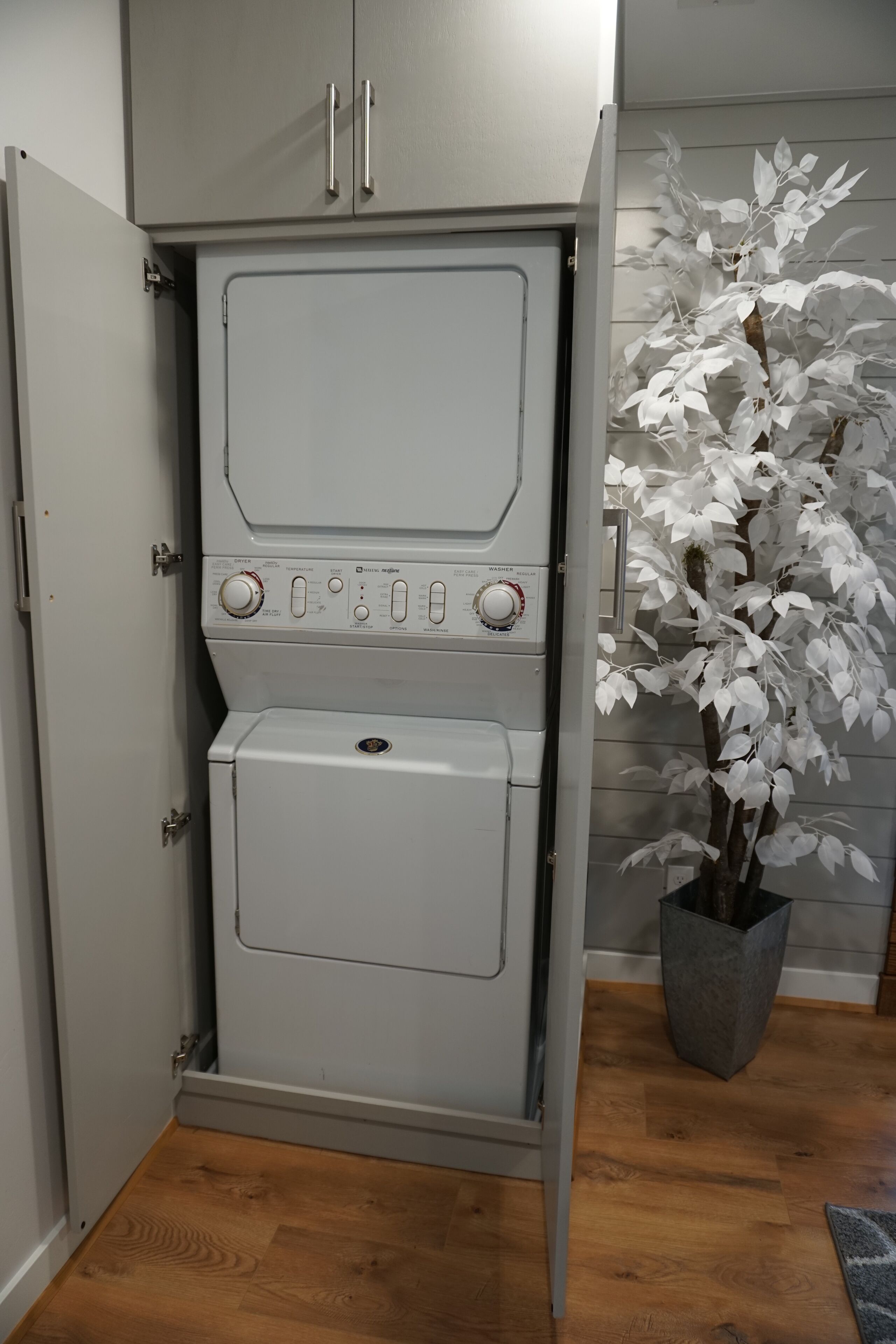 A compact, grey utility room with a built-in washer and dryer, a potted plant, and a wooden floor.