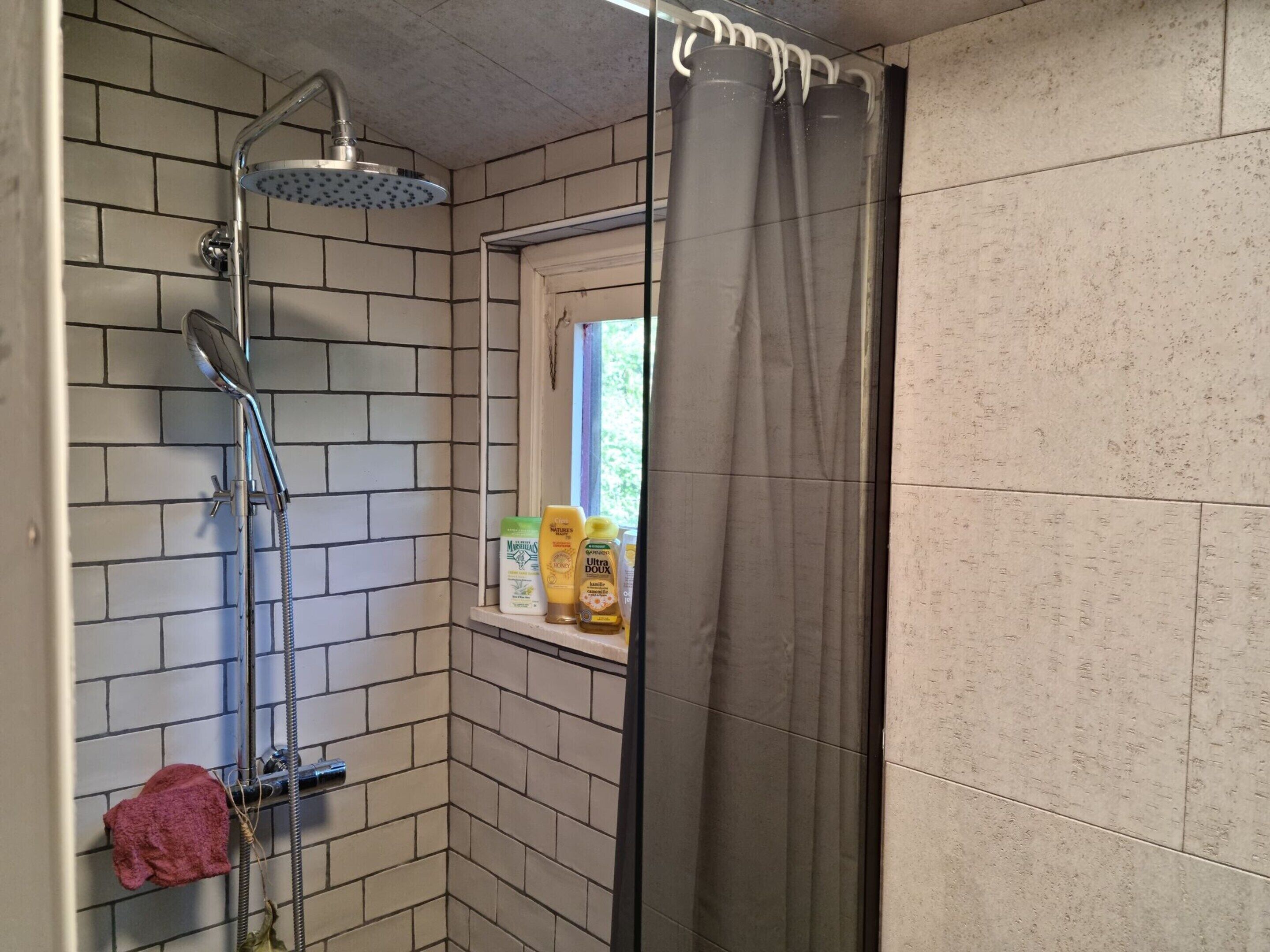A shower with a rain showerhead, a window with a view, and toiletries on a shelf.
