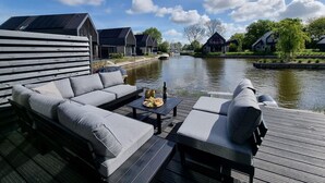Ein Uferdeck mit einer Couch und einem Tisch mit Blick auf einen Kanal, gesäumt von Häusern und Bäumen.