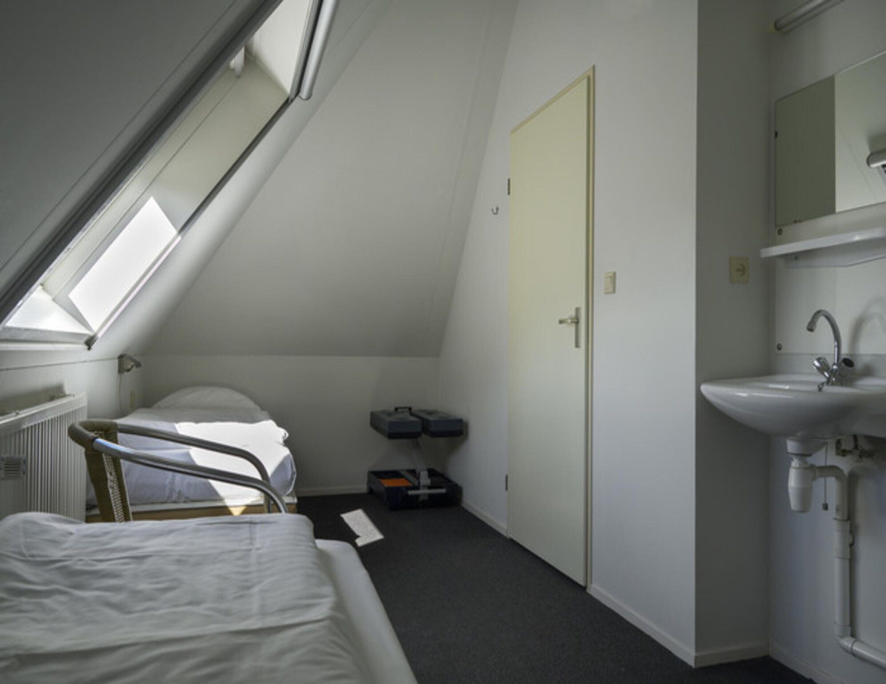 Una habitación pequeña y poco iluminada con una cama individual, una puerta, un radiador y un lavamanos sencillo.