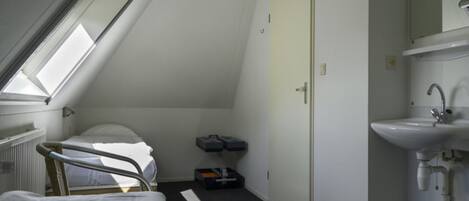 Una habitación pequeña y poco iluminada con una cama individual, una puerta, un radiador y un lavamanos sencillo.