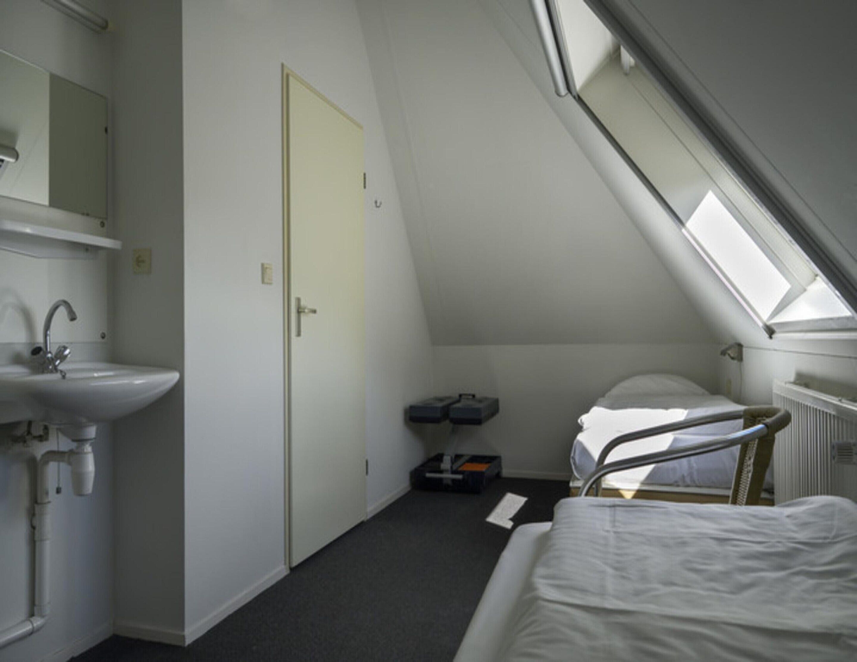 Une petite chambre avec un lit simple, dotée d’une lucarne, d’une table de chevet, d’un radiateur et d’une salle de bain avec un lavabo et un miroir.