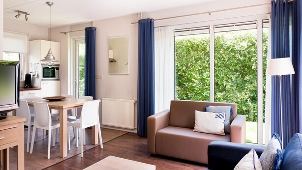 A modern living room with a sofa, coffee table, and dining area. There is a kitchen with a microwave and a TV. Large windows with curtains provide a view of greenery outside.