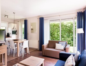 Un salon moderne comprenant un canapé, une table basse et un coin repas. Il y a une cuisine équipée d’un four à micro-ondes et d’un téléviseur. De grandes fenêtres, ornées de rideaux, offrent une vue sur la verdure environnante.
