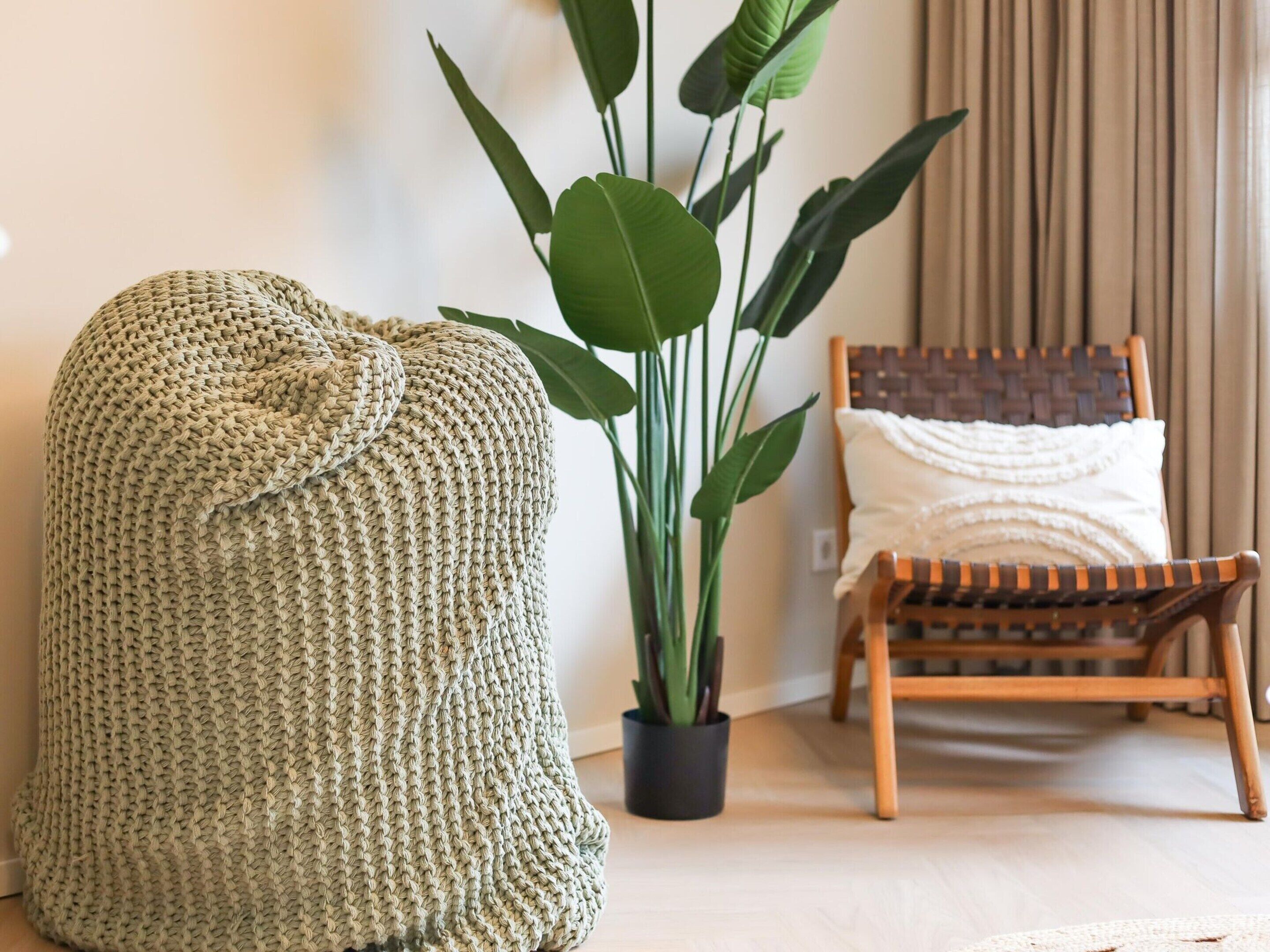 Une pièce comprenant un grand pouf tissé, une plante en pot, une chaise en bois et un coussin blanc.