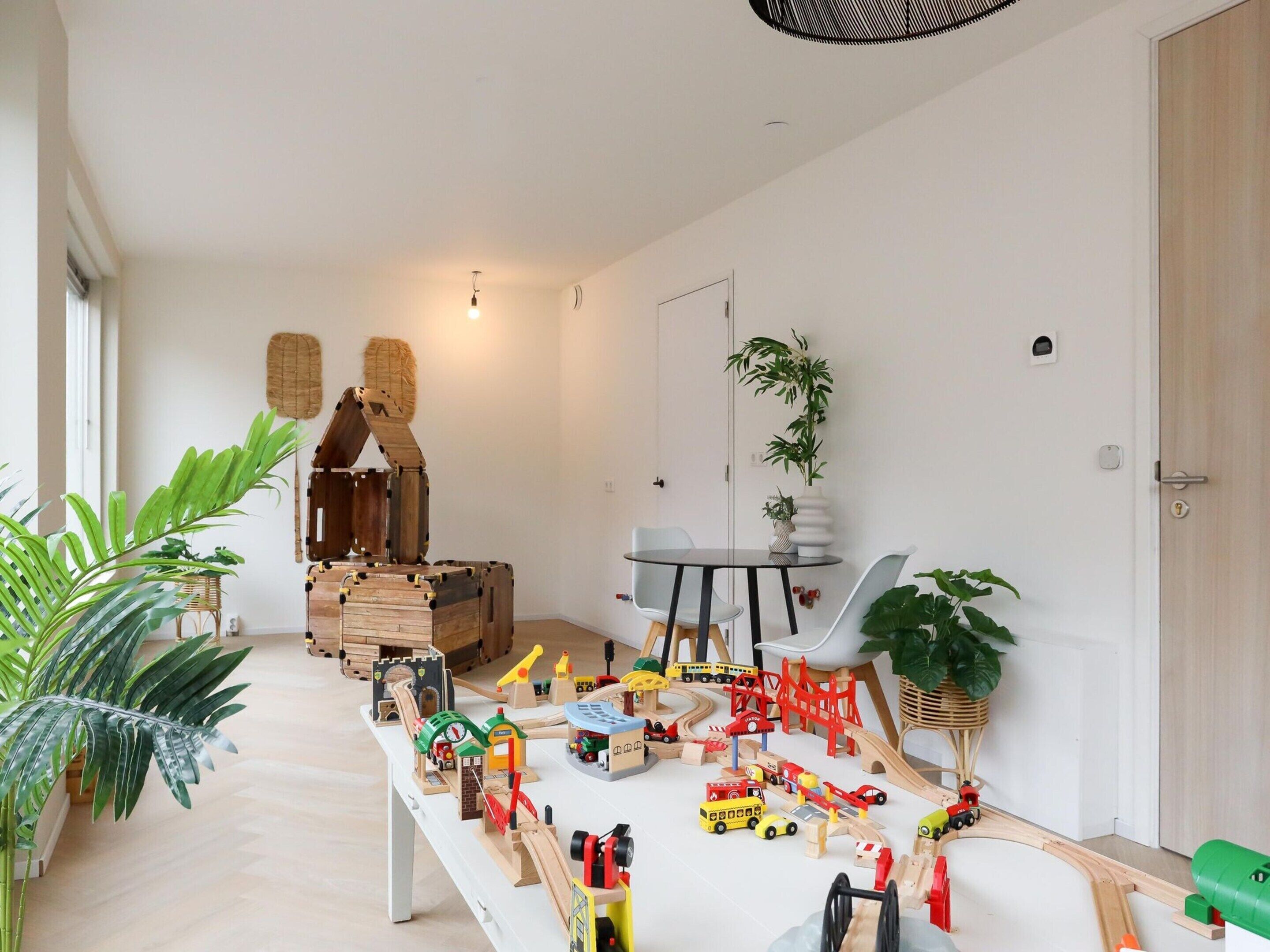 A room with a play area featuring wooden train tracks and toy vehicles. There are potted plants and a dining table with chairs.