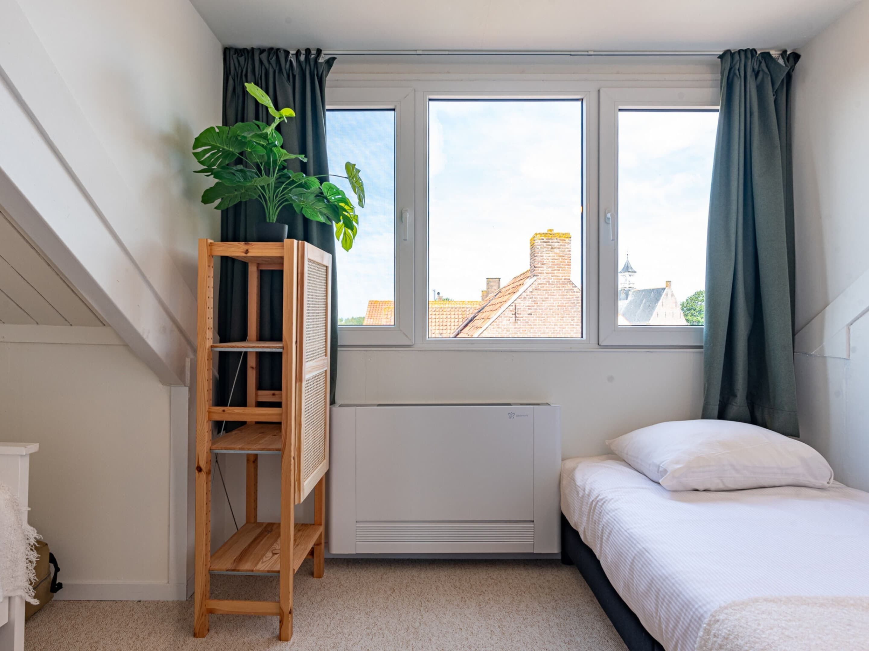 A bedroom with a bed, a radiator, a window with curtains, and a wooden ladder with a plant.