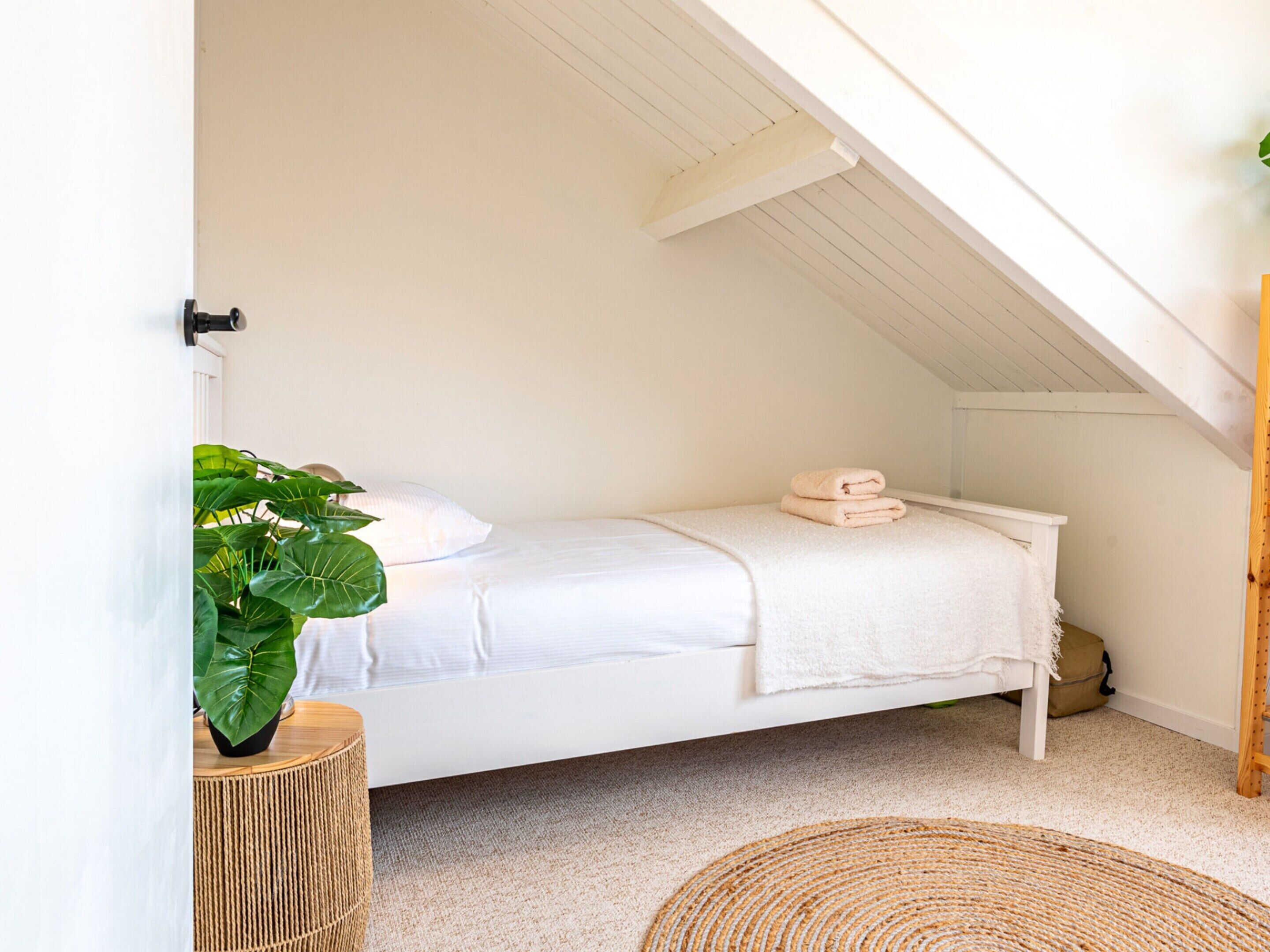 Un lit blanc dans une pièce dont le plafond est en pente, avec une plante et un tapis en fibres naturelles sur le sol.