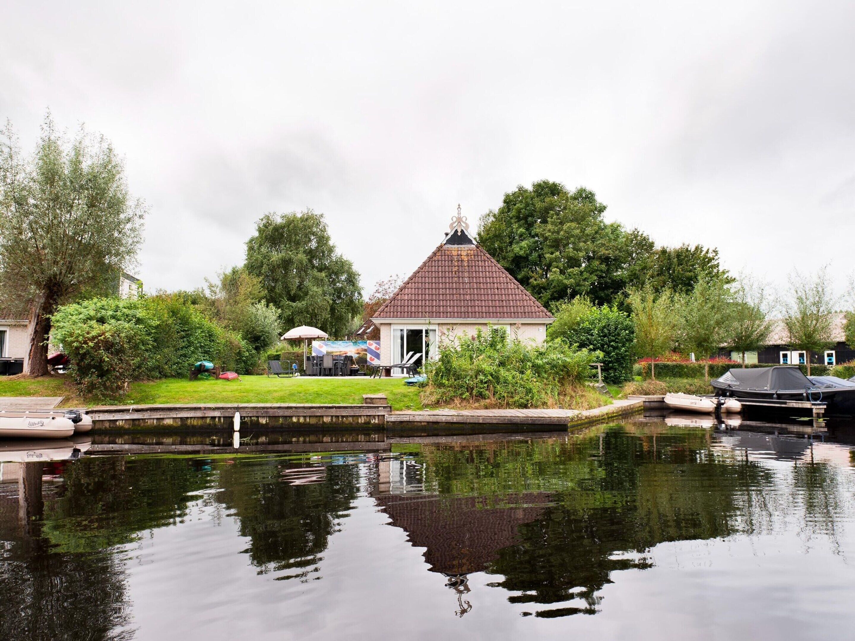 Une maison située au bord du canal, avec un toit en tuiles, un petit jardin et un quai où sont amarrés des bateaux.