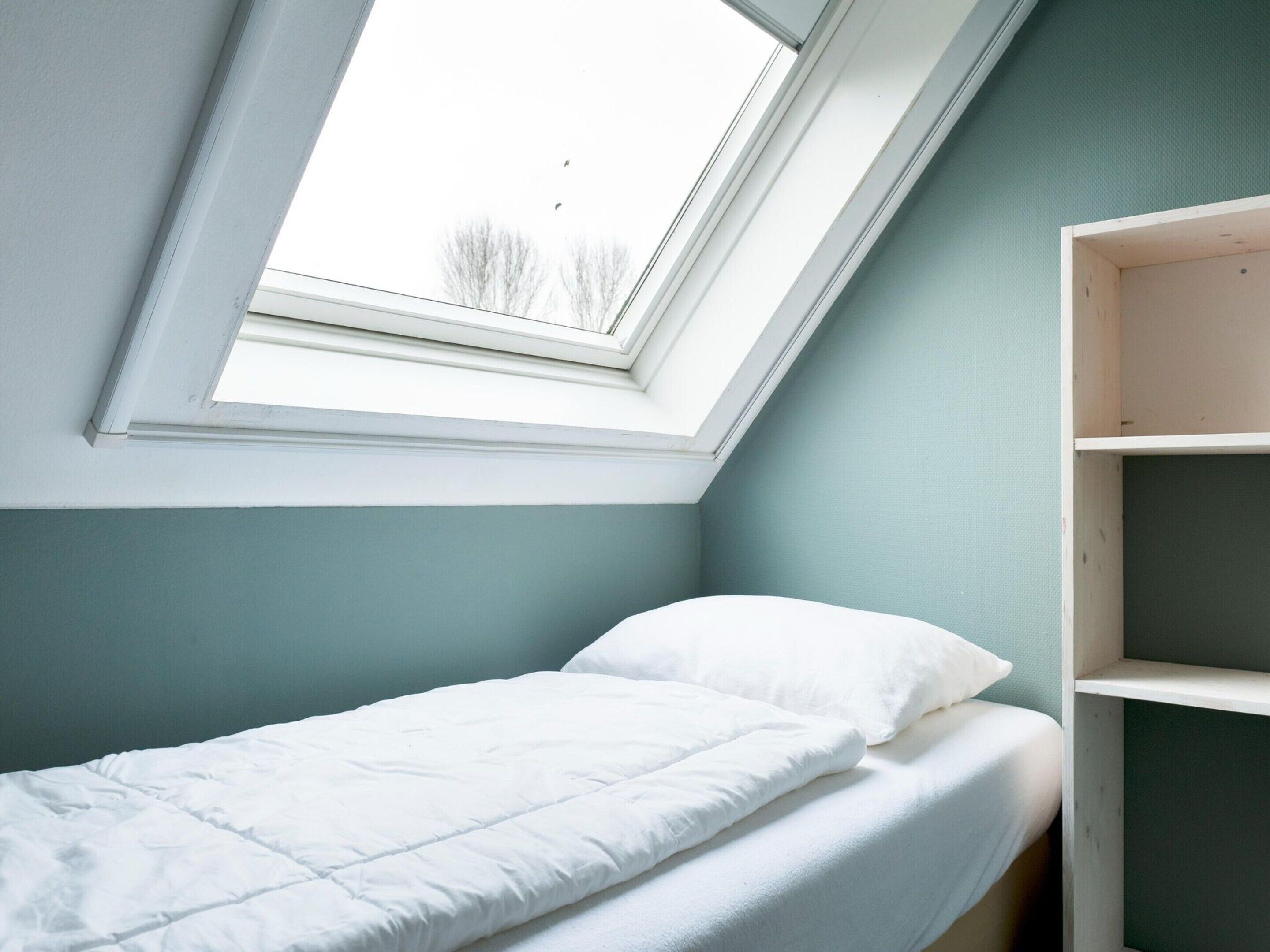 Quarto pequeno com cama de solteiro, claraboia, cama branca e estante de madeira.