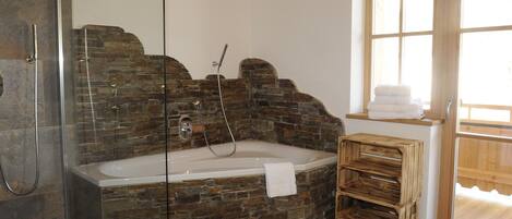 A bathroom with a stone wall, a glass-enclosed shower, a wooden bench, and a window with towels.
