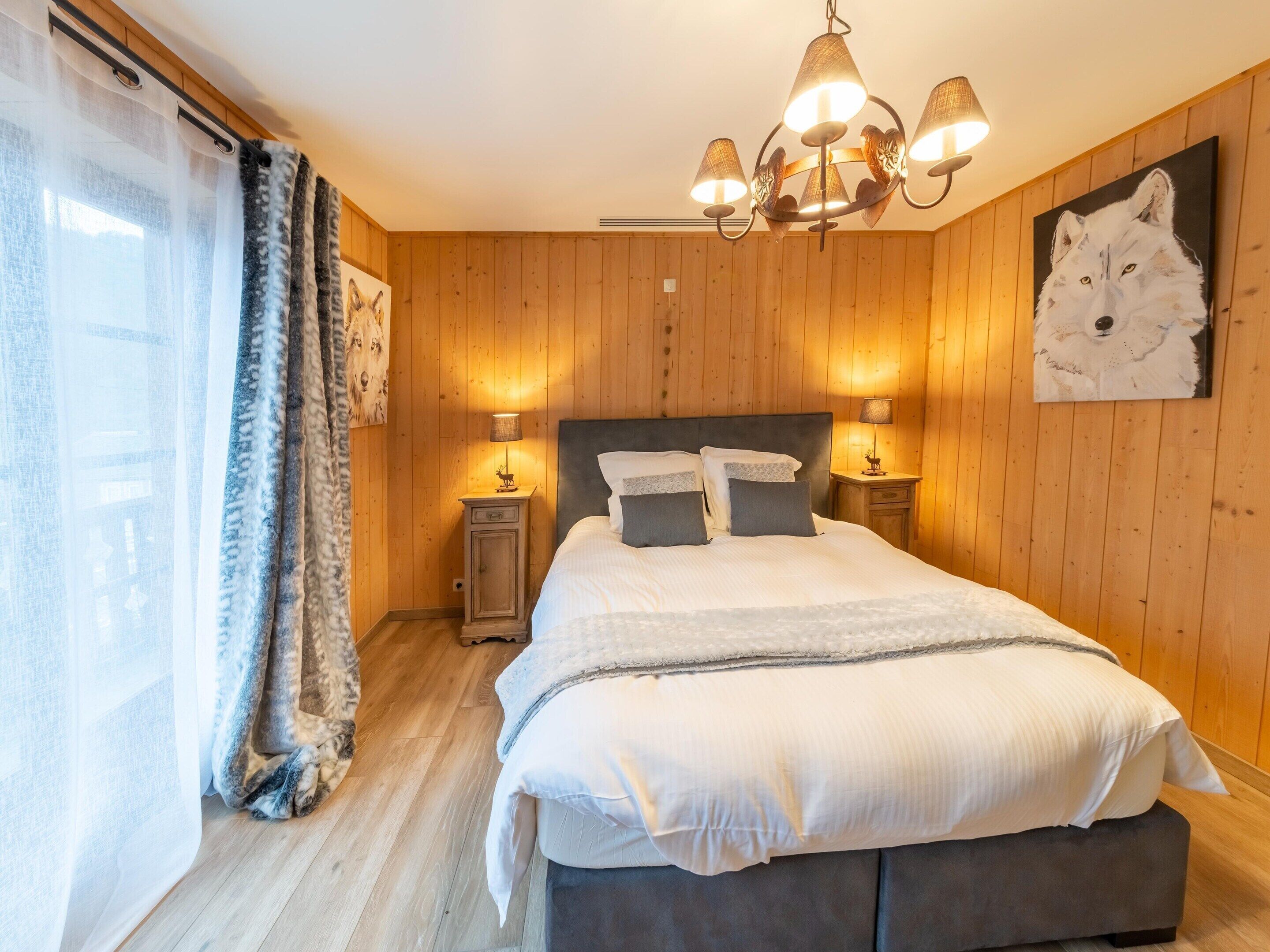 A bedroom with a wooden paneled wall, a bed with white bedding, a chandelier, and a painting of a wolf.