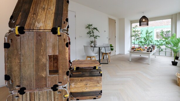 A modern interior with a wooden structure resembling a stack of suitcases, a dining area with a table and chairs, and a view of greenery through a window. - Shared Garden Apartment Retreat (Vrouwenpolder)