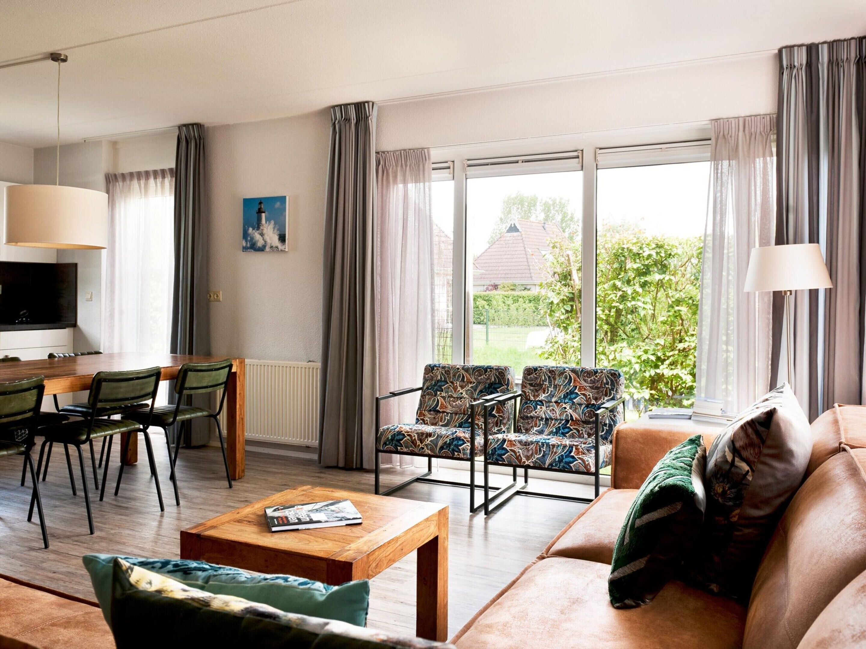 A modern living room with a sofa, two patterned armchairs, a wooden coffee table, a dining area with a table and chairs, and large windows with curtains.