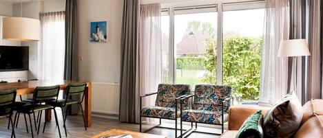Una sala moderna con un sofá, dos sillones estampados, una mesa de centro de madera, un comedor con mesa y sillas, y ventanas amplias con cortinas.