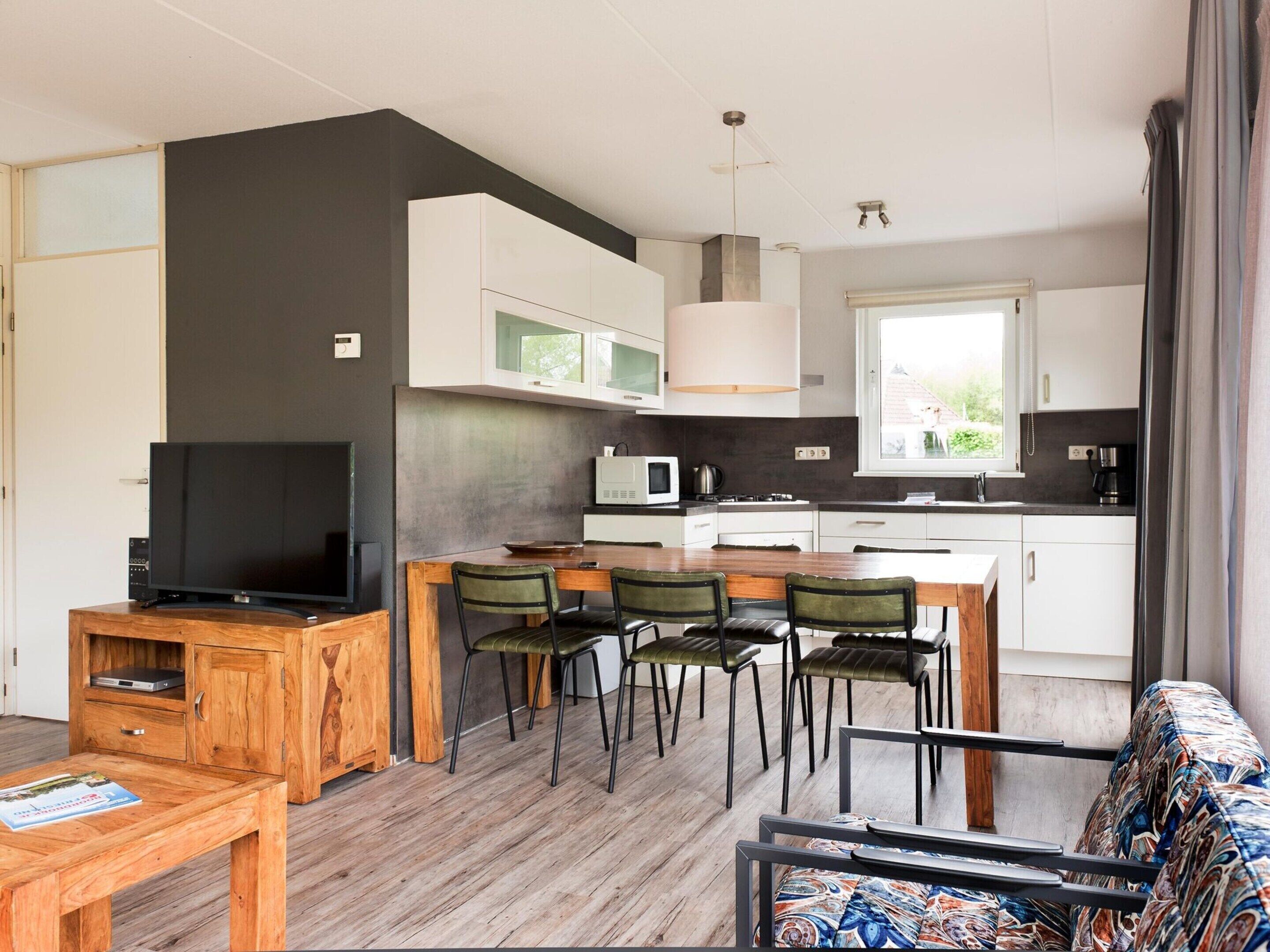A modern kitchen with white cabinets, a dining area with wooden tables and chairs, a television, and a window with curtains.