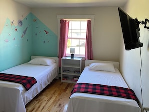Two single beds with checkered blankets, a flat-screen TV, and a nightstand with a lamp and books.