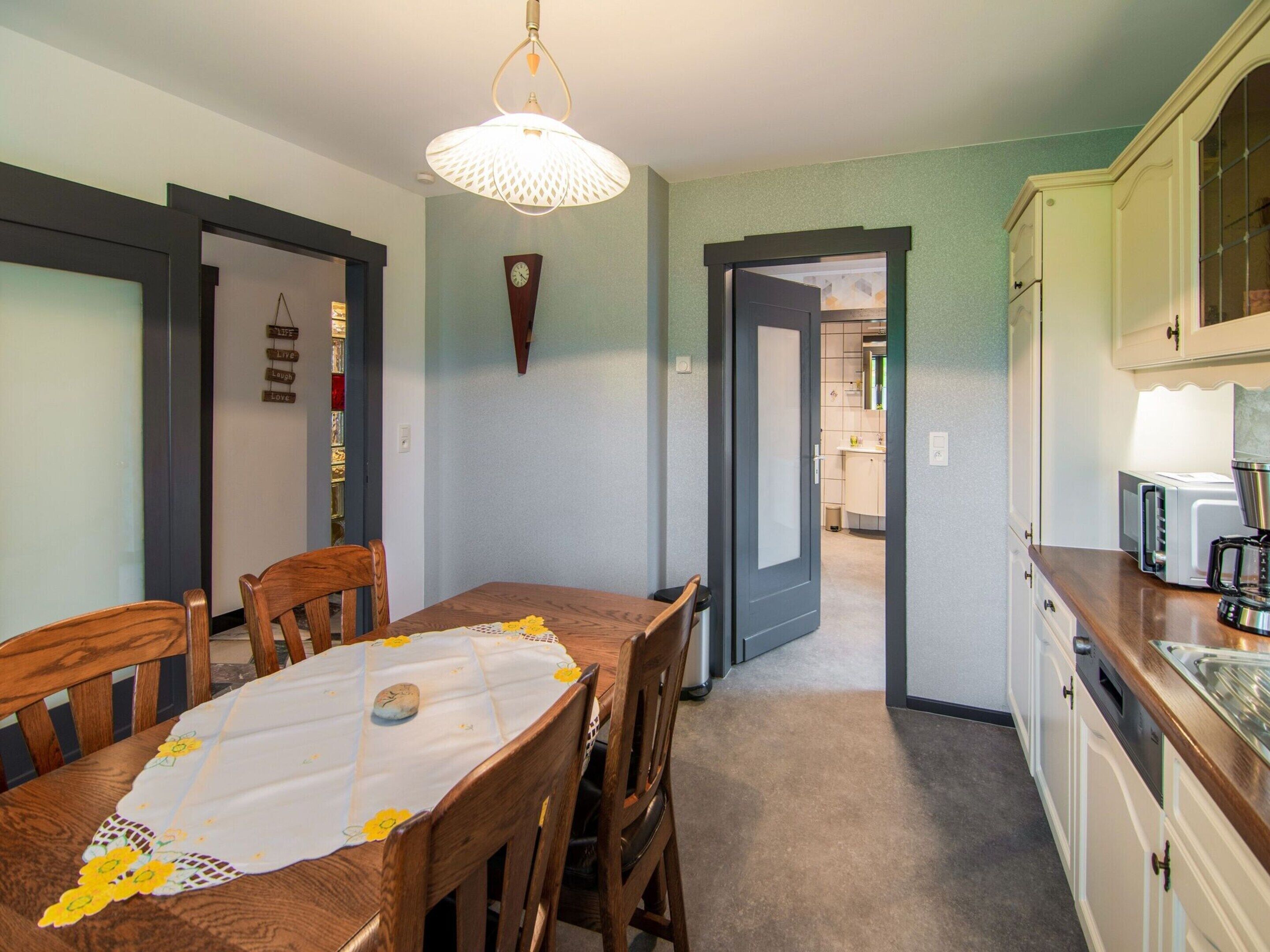 Een eetruimte met een houten tafel en stoelen, een decoratieve wandklok, en een keuken met witte kasten en een koffiezetapparaat.
