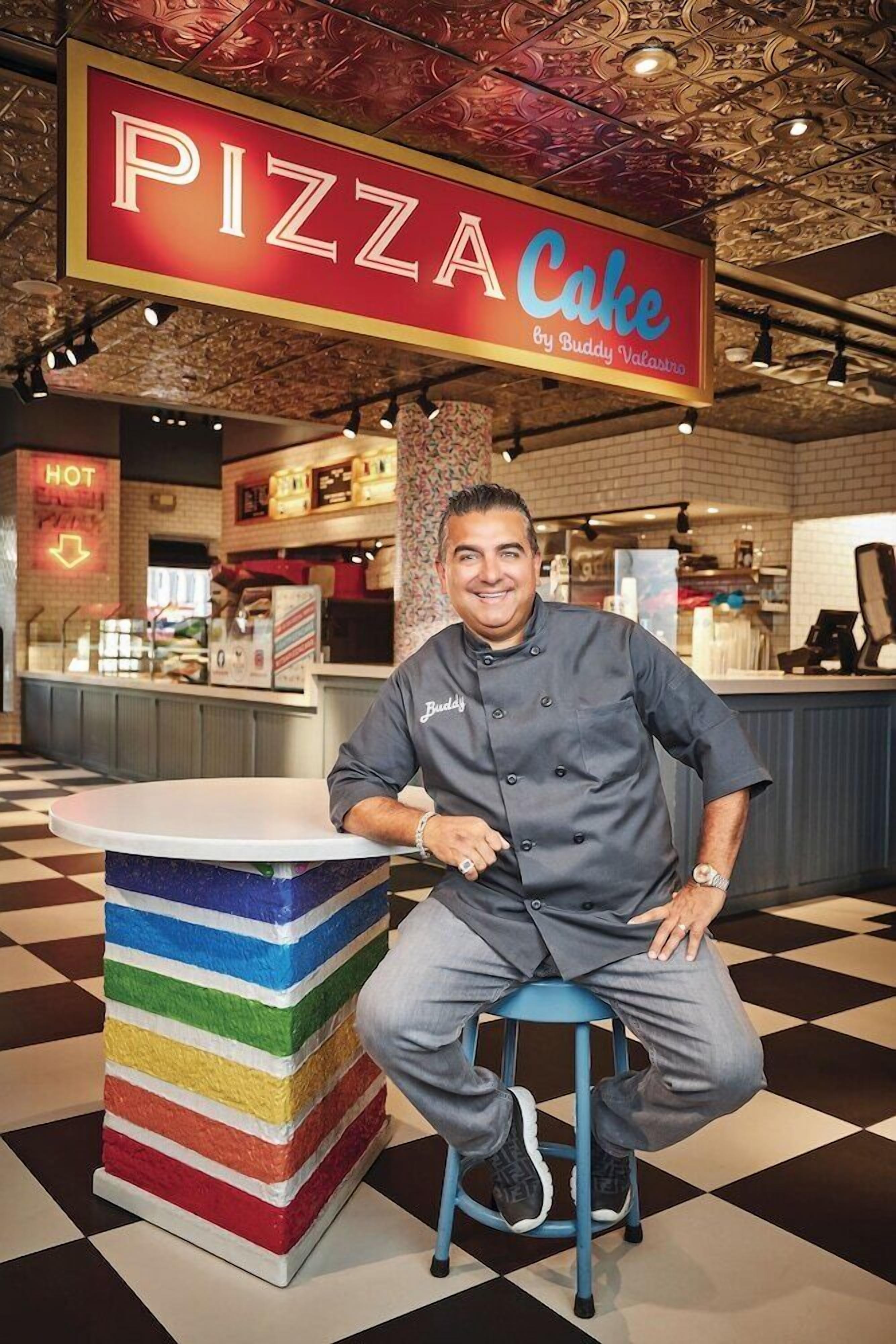 A chef seated at a colorful, multi-tiered stool in a restaurant with a 'PIZZA Cake' sign above.