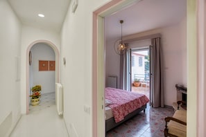 Un dormitorio con cama, un radiador, una ventana con cortinas, una puerta y un pasillo con radiador y plantas en macetas.