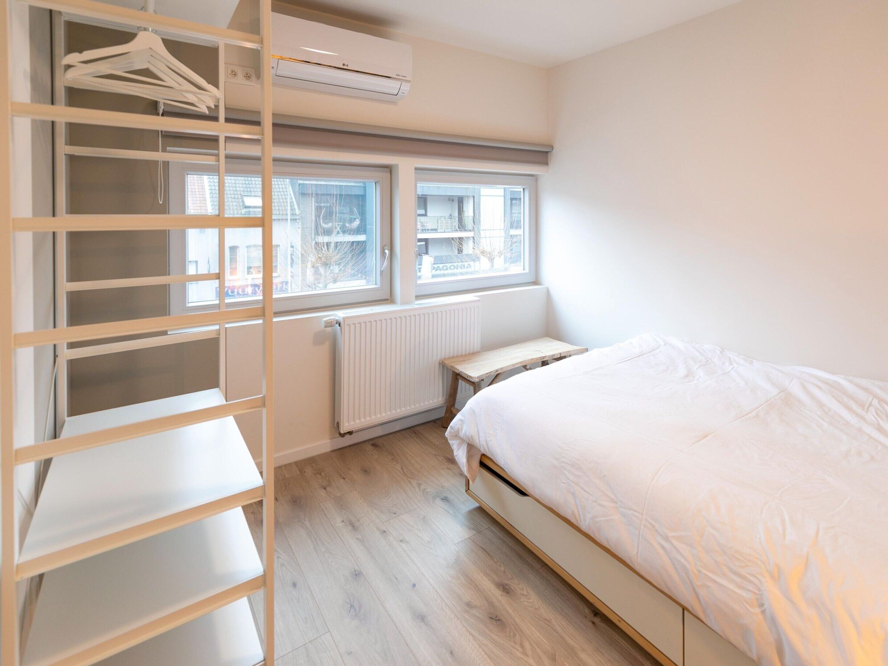 Une chambre à coucher avec un lit, une armoire en bois, un radiateur et une fenêtre donnant sur des immeubles.