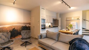 Sala de estar moderna com sofá, poltrona e mesa de centro. Cozinha visível ao fundo, com área de refeições.