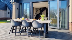 Area esterna con zona pranzo, tavolo e sedie, una struttura in vetro e vista sull'interno dell'edificio.