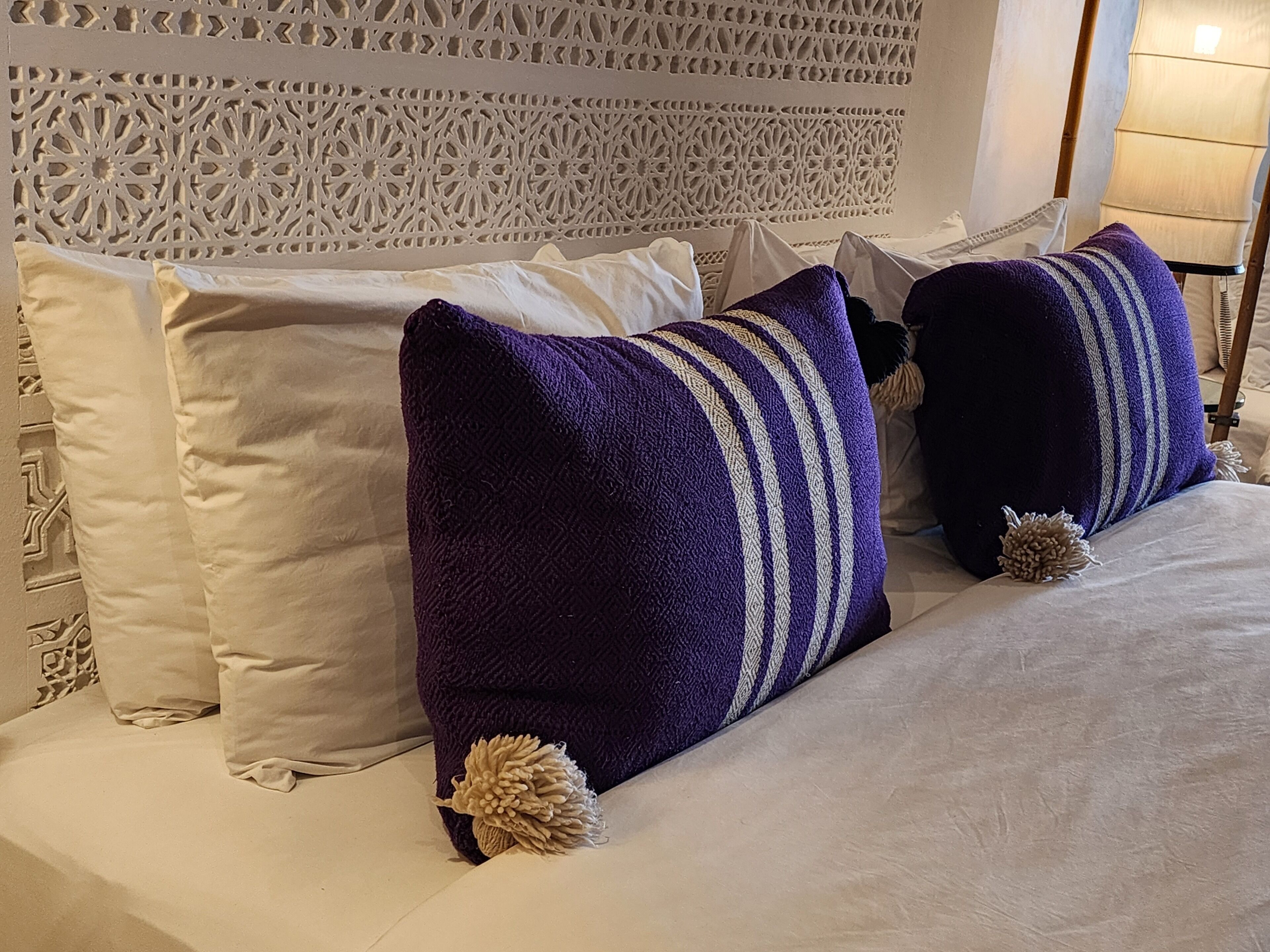 Een bed met witte en paarse kussens, een decoratief hoofdbord en een lamp.
